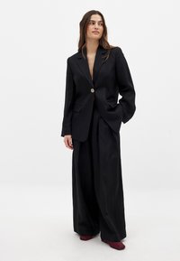 Melna pārspīlēta bleidere ar teksturētu apdari, ar vienu pogu un divām priekšējām kabatām, apvienota ar platām melnām biksēm un bordo kurpēm.