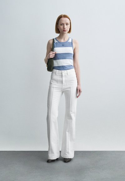 Ριγέ μπλε και λευκό ριμπ τοπ χωρίς μανίκια συνδυασμένο με λευκά τζιν παντελόνια με ψηλή μέση και φαρδύ καμπάνα; η μοντέλο κρατά μια μικρή πράσινη τσάντα, στέκεται σε γκρι πάτωμα.