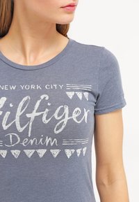 Camiseta de algodón azul con cuello redondo que presenta un estampado gráfico blanco que dice "NEW YORK CITY Hilfiger Denim" y triángulos decorativos.