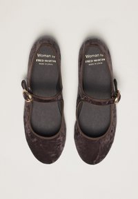 Un paio di scarpe Mary Jane in velluto marrone scuro con cinturini a fibbia dorata, etichettate "Woman by Fred Martin, made in Spain", su uno sfondo chiaro.