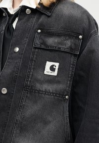 Άτομο φοράει μαύρο denim μπουφάν Carhartt με ασημί κουμπιά πάνω από λευκό πουκάμισο και μαύρη γραβάτα, εμφανίζοντας κοντινή λήψη της τσέπης του μπουφάν και του λογότυπου.