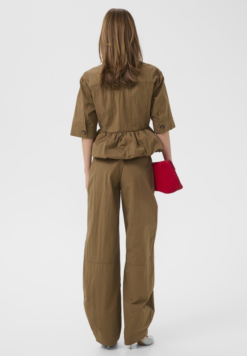 Vrouw met lang haar draagt bruine losse broek en jas met een getailleerde taille, houdt een felrode clutch vast en staat tegen een witte achtergrond.