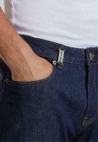 Jeans de mezclilla azul oscuro con costuras amarillas, un diseño sutil de bolsillo y un cierre de botón metálico, que cuenta con una pequeña etiqueta de marca en la cintura.
