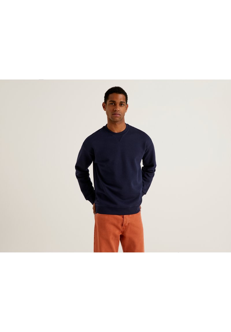 Hombre vestido con un sudadera de manga larga azul marino y pantalones naranjas, de pie con las manos detrás de la espalda frente a un fondo liso.