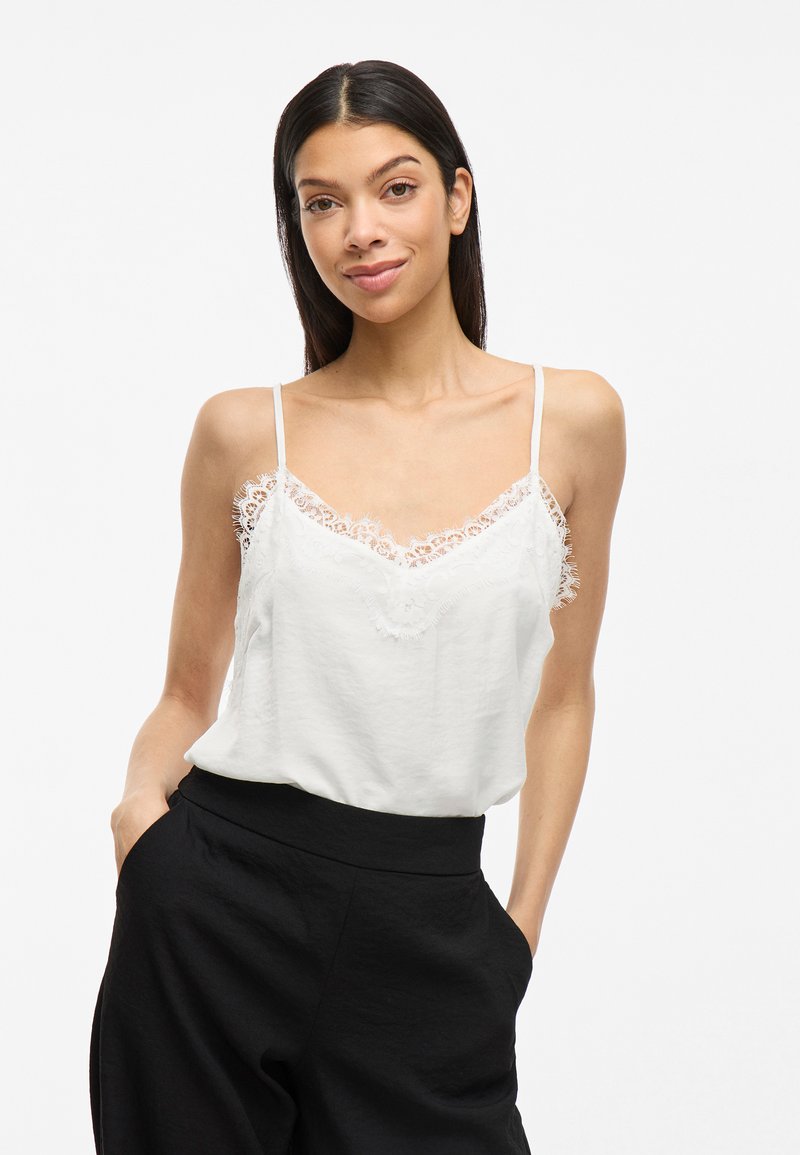 Camiseta blanca sin mangas con detalles de encaje en el escote y tirantes finos, combinada con pantalones de tiro alto negros, diseño minimalista.