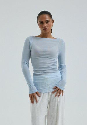 Frau mit zurückgebundenem Haar, die ein hellblaues, eng anliegendes Langarmshirt und lockere weiße, gemusterte Hose vor schlichtem Hintergrund trägt.