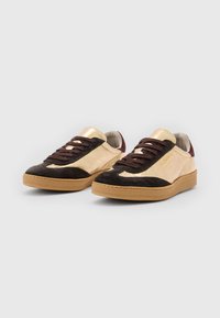 Sneakers in suede oro e marrone con lacci scuri e suola in gomma. Presentano dettagli metallici vicino al tallone e un logo sul lato.
