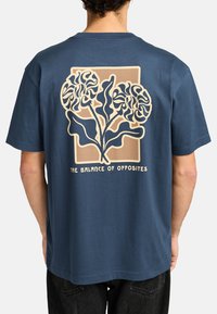 Navyblaue Baumwoll-T-Shirt mit einem großen, abstrakten Blumenmotiv auf der Rückseite in Beige, darunter der Text "DAS GLEICHE DER GEGENSÄTZE".