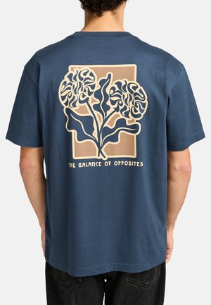 Marineblauwe katoenen t-shirt met een groot, abstract bloemenmotief op de achterkant in beige, met de tekst "DE BALANS VAN TEGENSTELLINGEN" eronder.