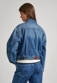 Veste en denim bleu moyen, design court avec une taille en maille blanche côtelée. Présente des coutures classiques et une patch en cuir marron au dos.
