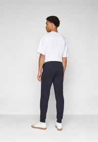 T-shirt bianco a maniche corte con vestibilità ampia, abbinato a pantaloni sportivi blu scuro con polsini a costine e sneakers bianche con suola beige.