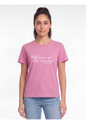 Junge Frau mit dunklem, zurückgebundenem Haar, die ein pinkes T-Shirt mit deutschem Text und hellblaue Jeans vor weißem Hintergrund trägt.