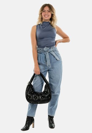 Glunderende vrouw in een mouwloos blauw topje, hooggewaaid knielange lichte jeans, zwarte enkellaarzen, met een zwarte handtas met studs aan de zijkant.