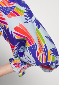 Bras portant un tissu bleu clair avec des motifs abstraits de coups de pinceau colorés en violet, rouge, orange, rose et vert, froncés au poignet.