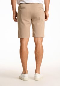 Pantaloni corti beige con una texture liscia, vestibilità slim e orli arrotolati. Presentano due tasche posteriori con botttoni e sono abbinati a scarpe da ginnastica bianche.