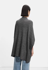 Kullgrå strikket poncho med brede ermer og rund fald, båret over lyseblå vide bukser. Glatt tekstur og minimalistisk design.