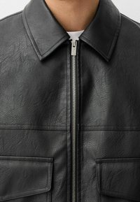 Zwarte neplederen jas met een puntige kraag, ritssluiting aan de voorkant en twee voorzakken. Gerenoveerd materiaal met zichtbare stikdetails.