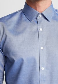 Muž oblečený v světle modré texturované košili na knoflíky s špičatým límcem, odhalující horní část hrudníku a krku, žádné další detaily nejsou viditelné.