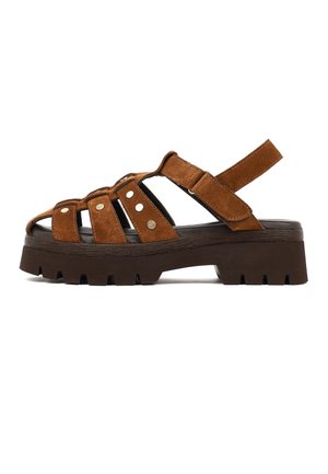 Sandale en suède marron avec une semelle noire épaisse, bout ouvert, trois clous métalliques sur les brides et une bride de cheville réglable en Velcro.