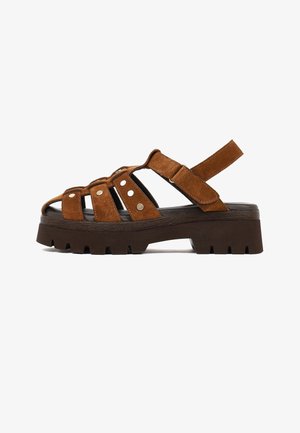 Sandalo in suede marrone con suola spessa nera, punta aperta, tre borchie metalliche sulle cinghie e cinturino alla caviglia regolabile in Velcro.
