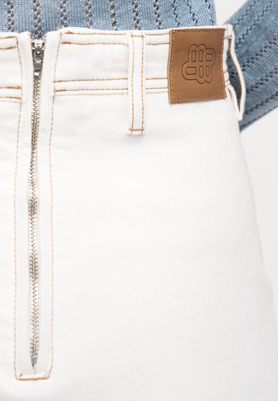 Pantalon en denim blanc avec une fermeture éclair à l'avant, des coutures orange contrastantes et une écusson en cuir marron sur la ceinture.