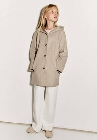 Beige vlnený kabát s kapucí, se střih na rovno, velkými knoflíky a postranními kapsami. Kombinovaný s bílými širokými kalhotami a béžovými pantoflemi.