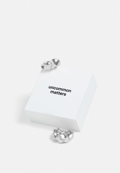Silvrigt vridet metallsmycke placerat bredvid en vit fyrkantig låda märkta "uncommon matters", med rena linjer och en slät yta.