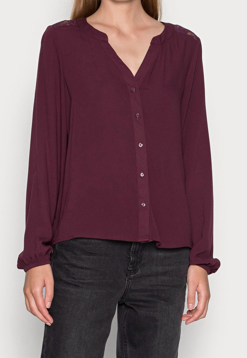 Blouse bordeaux à manches longues en tissu léger, avec un col en V, une fermeture à boutons sur le devant et des accents en dentelle sur les épaules.