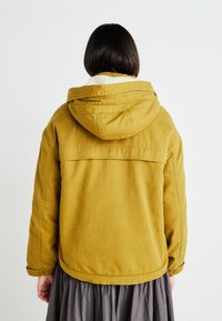 Geel hoodie-jack met een zachte, crèmekleurige kraag. Heeft elastische manchetten en een eenvoudige, onversierde achterzijde.