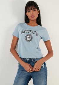 Gaiši zila īsa t-krekla ar apaļu kakla izgriezumu, īsām piedurknēm un melnu "BROOKLYN" tekstu un emblēmu uz priekšpuses.
