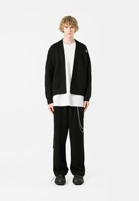 Cardigan negro con textura y diseño abierto, combinado con una camiseta blanca, pantalones negros holgados y calzado negro grueso con un patrón.