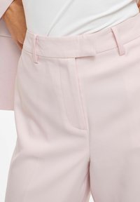 Pantalon rose clair avec une texture lisse, présentant un devant plat, des poches latérales et un détail subtil à la ceinture.