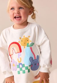 Sweat-shirt blanc orné d'appliqués colorés représentant un arc-en-ciel, un soleil, une poire, une fleur et un lapin. Col côtelé, manches ballon et design ludique.