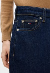 Los jeans de mezclilla azul oscuro presentan un diseño ajustado, costuras visibles, dos bolsillos delanteros con botones claros y un pequeño detalle de logo en rojo, blanco y azul.