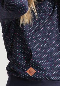 Marinblå sweatshirt med turkosa och rosa prickar, med en känguruficka och en läderlogotypsskylt vid nederdelen fram.