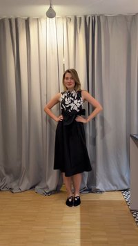 Γυναίκα στέκεται με τα χέρια στη μέση, φοράει αμάνικο μαύρο φόρεμα με λευκή φλοράλ μπλούζα και μαύρα φλατ παπούτσια, μπροστά από γκρι κουρτίνες.