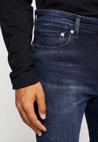 Mörkblå denimjeans med en slät textur, som har en framficka, två metallknappar och en kontrasterande midja. En svart långärmad tröja är delvis synlig.