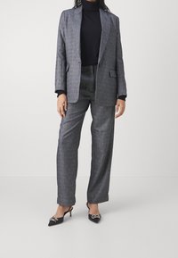 Costume à carreaux gris avec blazer sur mesure et pantalons à jambes larges, associé à un col roulé noir et des escarpins à bout pointu ornés d'un nœud décoratif.