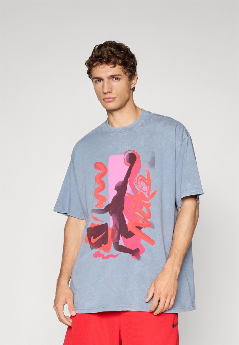 Ung mand iført en løs blå T-shirt med et rød- og pinkfarvet basketballgrafik og røde shorts foran en ensfarvet grå baggrund.