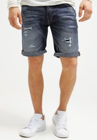 Person wearing rolled-up, distressed dark denim shorts and white sneakers, standing against a plain white background.