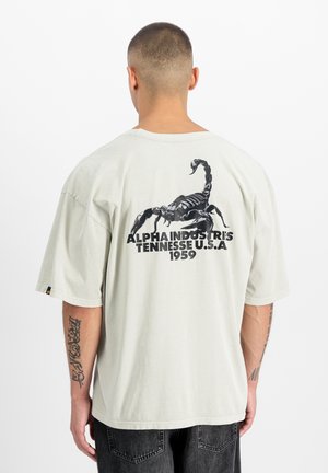Mann mit kurzen Haaren, von hinten gesehen, trägt ein hellbeiges T-Shirt mit einem großen schwarzen Skorpion und dem Text "Alpha Industries Tennessee U.S.A 1959."