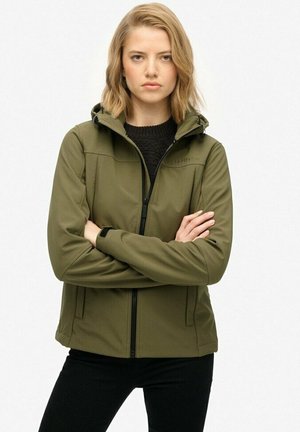 Jonge vrouw met blond haar die een olijfgroene gewatteerde jas en zwarte broek draagt, staande met de armen gekruist tegen een witte achtergrond.
