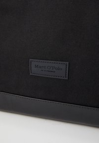 Black canvas bag with a smooth black leather bottom. Features a rectangular brand label embossed with "Marc O'Polo" and "EST. IN STOCKHOLM."