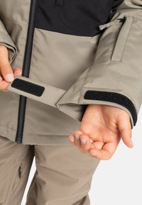 Osoba dostosowująca czarny rzep na mankiecie beżowej kurtki, ubraną w dopasowane beżowe spodnie, z widocznym zamkiem błyskawicznym z przodu kurtki.