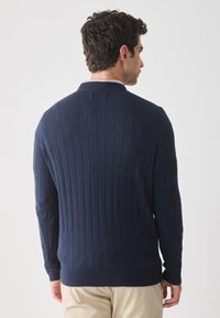 Homme de dos portant un pull en maille bleu marine et un pantalon beige sur un fond clair uni.