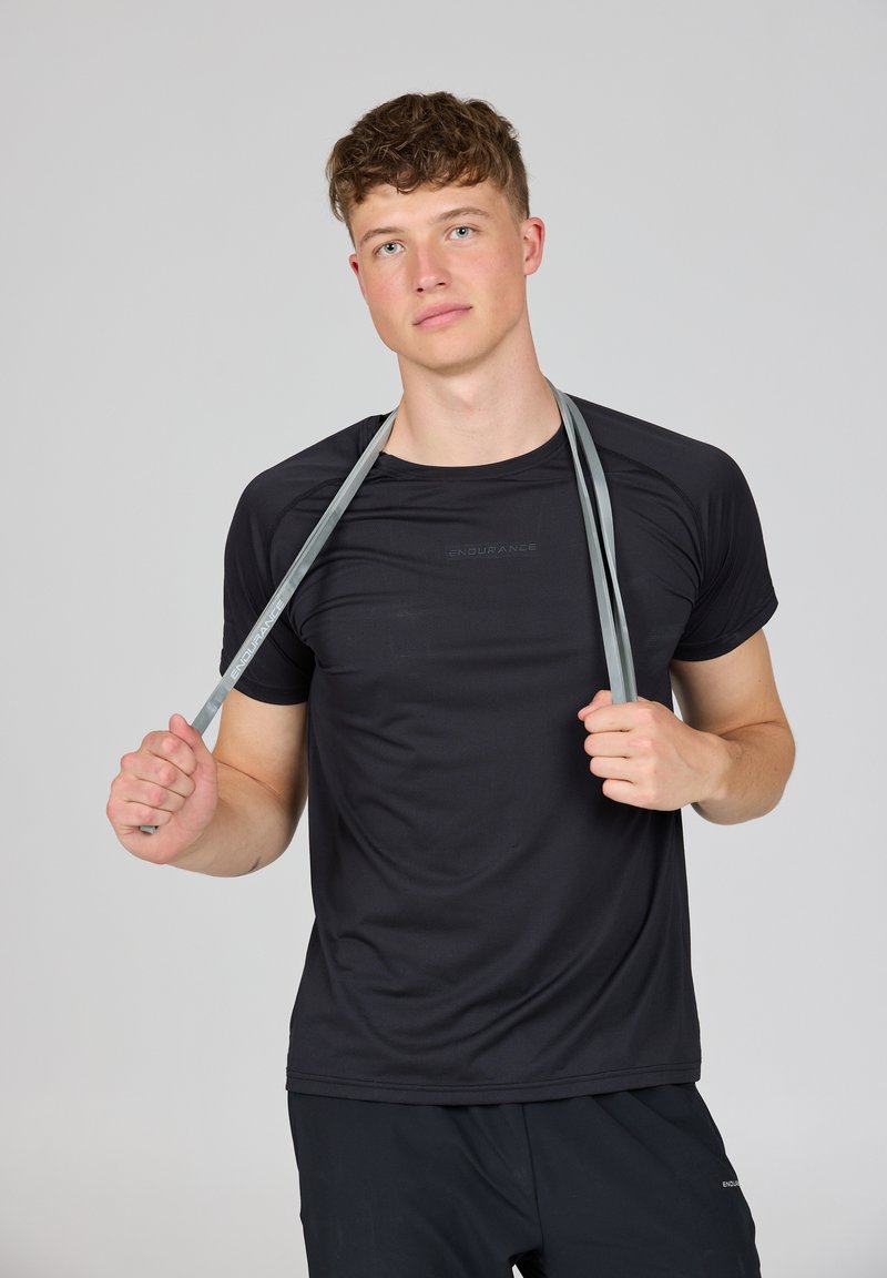 Jonge man in zwarte sportkleding die een grijze weerstandsband over zijn schouders houdt tegen een effen achtergrond.
