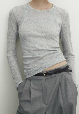 Persoon die een lichtgrijs aansluitend shirt met lange mouwen draagt over een bijpassende tanktop, ingestopt in hoge taille grijze geplooide broek met een zwarte riem.