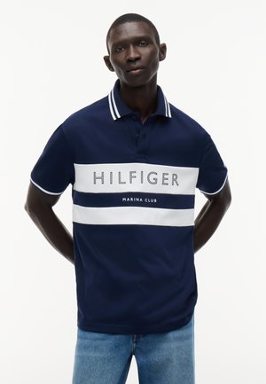 Mand iført en marineblå og hvidstribet polo med teksten "Hilfiger Marina Club", kombineret med lyseblå jeans, står med hænderne bag ryggen.