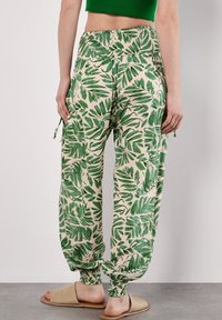 Une personne se tient debout, portant un pantalon à motifs de feuilles vertes et des chaussures ballerines beiges, avec un mur lisse en arrière-plan.