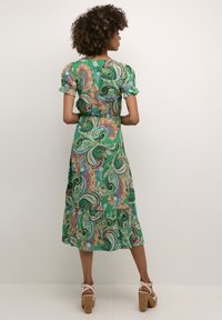 Green paisley patterned dress with short puff sleeves, a cinched waist, and tiered hem. Worn with beige strappy heels.